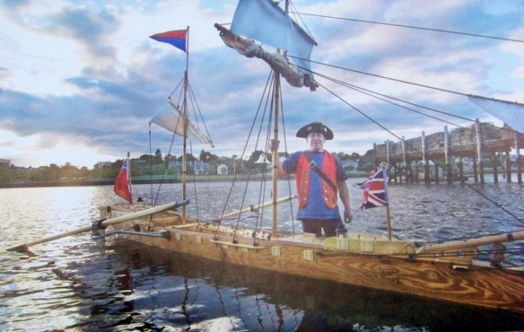 A person in historical costume stands on a wooden boat with flags on a calm waterfront.